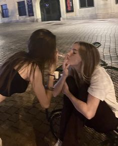 two young women sitting on a bench in front of a building, one biting the other's mouth