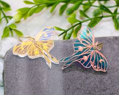 two butterfly brooches sitting on top of a gray rock next to green leaves