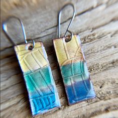 two square shaped earrings with blue and green designs on them sitting on a wooden surface