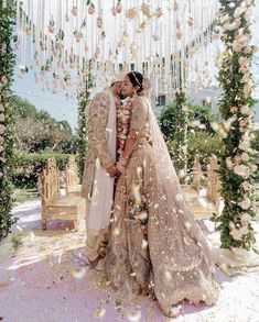 two people in wedding outfits standing under a flower covered arch with petals falling from the ceiling