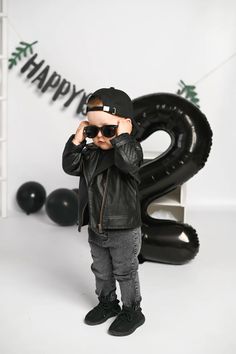 a young boy wearing sunglasses and a leather jacket, standing in front of a black balloon