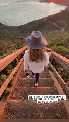 a woman wearing a hat walking up some stairs with the ocean in the back ground