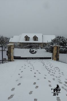 an iron gate with footprints leading to a house covered in snow and surrounded by trees