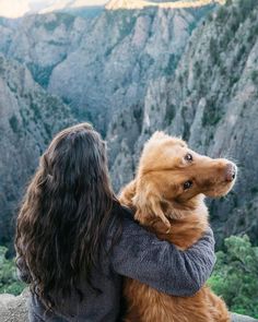 a woman holding a dog in her arms on top of a mountain with mountains in the background