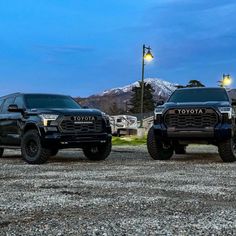 two black trucks parked next to each other in front of a mountain range at night