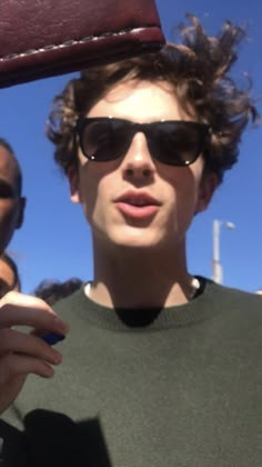 a young man wearing sunglasses and holding a cell phone up to his face with other people in the background