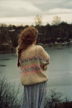 a woman standing on top of a hill next to a body of water wearing a sweater