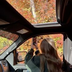 a woman is taking a photo in the rear view mirror of a car with autumn foliage behind her