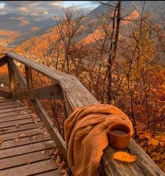 a blanket and cup sitting on top of a wooden bench in front of autumn foliage