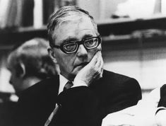 black and white photograph of a man in glasses sitting at a table with his hand on his chin