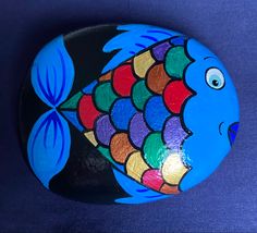 a painted rock with an image of a colorful fish on it's side, sitting on a blue surface