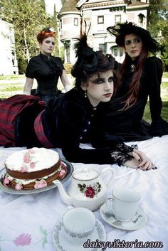 three women dressed in victorian clothing sitting at a table with a cake and tea cups