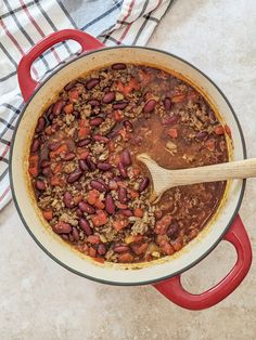 a red pot filled with chili and beans on top of a white table cloth next to a wooden spoon