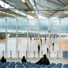 several people are playing basketball in an indoor court with blue chairs and large windows on the side wall