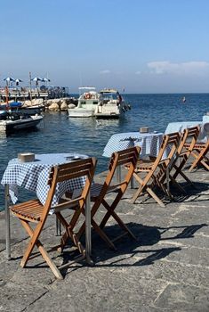 there are many chairs and tables along the water's edge with boats in the background