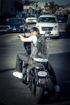 a man riding on the back of a motorcycle down a street next to parked cars