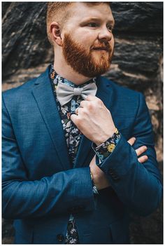 a man with a beard wearing a blue suit and bow tie standing in front of a rock wall