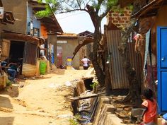 a woman sitting on the side of a dirt road next to a tree and buildings