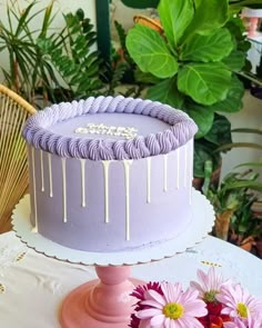 a purple cake with white icing sitting on top of a table next to flowers