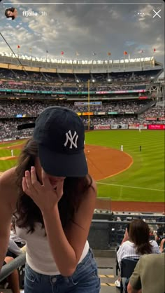 a woman in a yankees hat sitting at a baseball field while talking on her cell phone