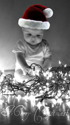 a black and white photo of a baby wearing a santa hat sitting in front of christmas lights