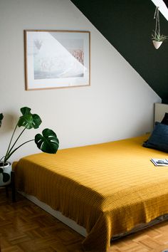 a bed with a yellow blanket in a room next to a plant and a painting on the wall