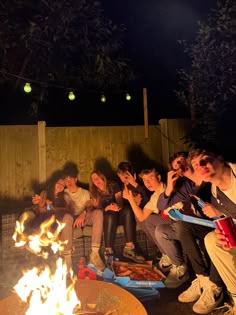 a group of people sitting around a fire pit at night with their hands in the air