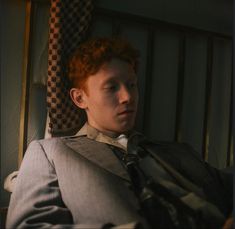 a man with red hair wearing a suit and tie sitting in a chair looking off to the side