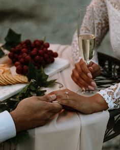 two people sitting at a table holding glasses of wine with grapes and cheese on the side