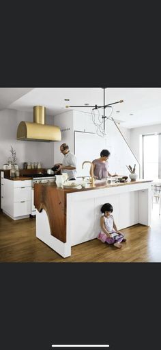 two people are cooking in the kitchen with white cabinets and wood flooring, while one person is sitting on the counter