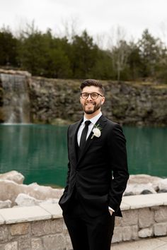 a man in a tuxedo standing next to a lake with a waterfall behind him