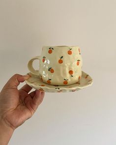 a hand holding a ceramic cup and saucer with oranges on the rim, in front of a white background