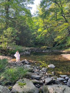 a woman standing on rocks in the middle of a river surrounded by trees and grass