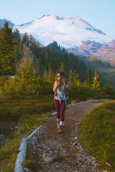 a woman walking down a trail in front of a snow covered mountain with trees on both sides