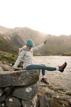 a woman sitting on top of a stone wall next to a lake with mountains in the background