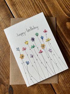 a happy birthday card with colorful flowers on the front and bottom, sitting on top of a wooden table