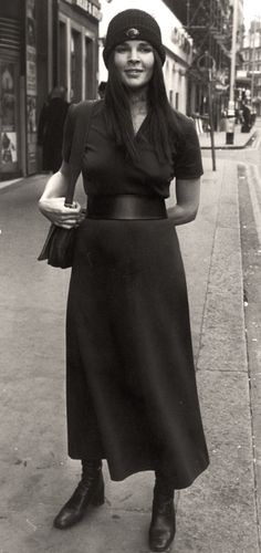 a woman is standing on the sidewalk with her purse in hand and wearing a hat