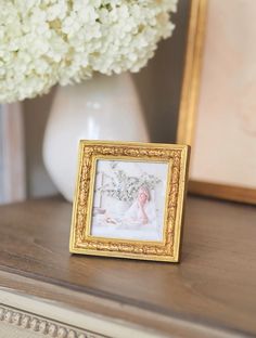 a gold frame sitting on top of a wooden table next to a vase with white flowers