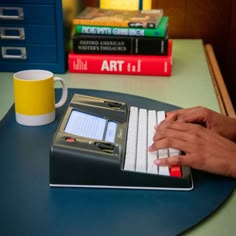 a person typing on a computer keyboard next to some books and a cup with a yellow mug