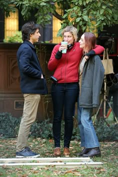 three people standing in front of a tree talking to each other and drinking from cups