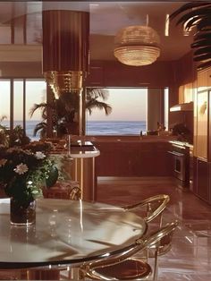 a dining room table surrounded by chairs and a vase with flowers on it in front of a large window overlooking the ocean