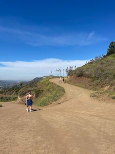 two people running down a dirt road on top of a hill with palm trees in the background