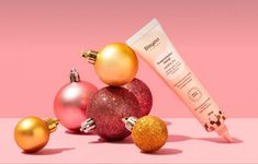 three christmas ornaments and a tube of sunscreen on a pink background with gold baubies