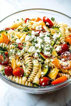 a pasta salad with tomatoes, zucchini and feta cheese in a glass bowl