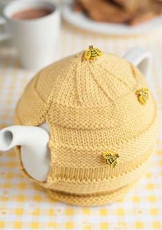 a knitted tea pot cover with two bees on it and a cup of coffee in the background