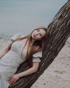 a woman in a white dress leaning against a tree on the beach with her eyes closed