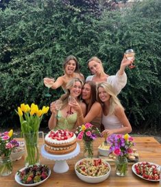 four women standing around a table full of food and drinks, posing for the camera