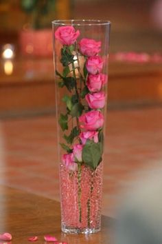 some pink roses in a clear vase on a wooden table with petals scattered around it