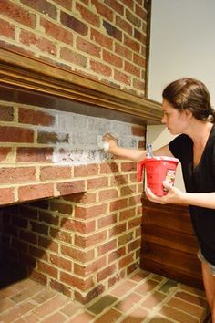 a woman is painting a brick fireplace with a red bucket and white paint on it