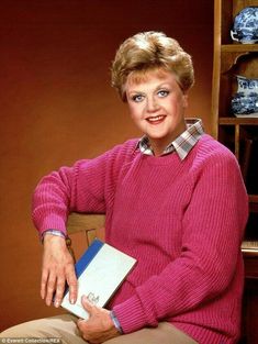 an older woman sitting in a chair holding a book and posing for the camera with her hands on her hips